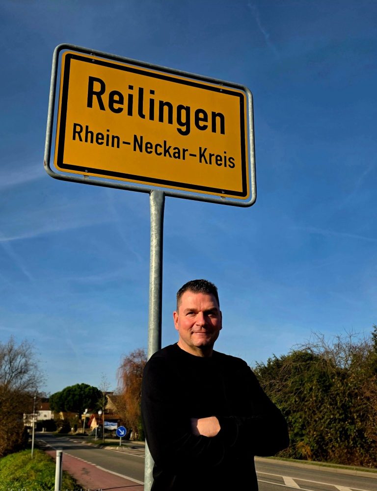 Schild mit "Reilingen" und Ortsschild, André Pottkamp steht daneben vor blauem Himmel.