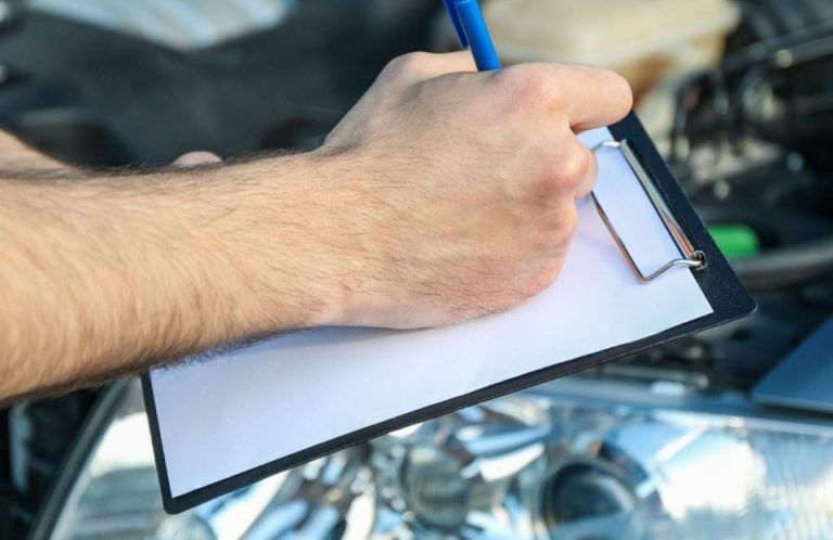 Hände halten einen Stift und eine Klemmtasche über dem Motorraum eines Fahrzeugs.